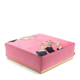 Square pink velvet pouf with a smooth gold base and print of hands applying lipstick on the top surface, viewed at an angle.