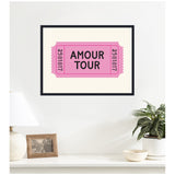 Framed pink retro ticket print with text 'AMOUR TOUR' and black numbers, hanging on a white wall above a minimalist white shelf with a lamp, photo frame, and plant.