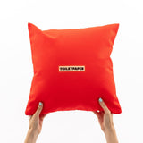 Bright red square pillow held up by two hands with light blue nail polish, featuring a small beige patch with black text reading 'TOILETPAPER' at the center.