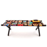 Bench with black wooden legs and a multicolored fabric seat covered in various rectangular and oval framed motifs, topped with a matching pillow on one end.