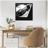 Black and white close-up photo of a vinyl record player’s turntable and tonearm, framed in black and hanging on a white wall above a wooden desk with beige chair.