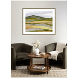 Abstract landscape painting with layered green, yellow, and gray horizontal bands framed in natural wood, hung above a cozy seating area with two dark armchairs and round wooden table.
