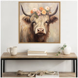 Framed painting of a Highland cow with long beige fur and large dark horns adorned with a crown of pink and orange flowers, hanging above a wooden table with books, a mug, and a white vase.