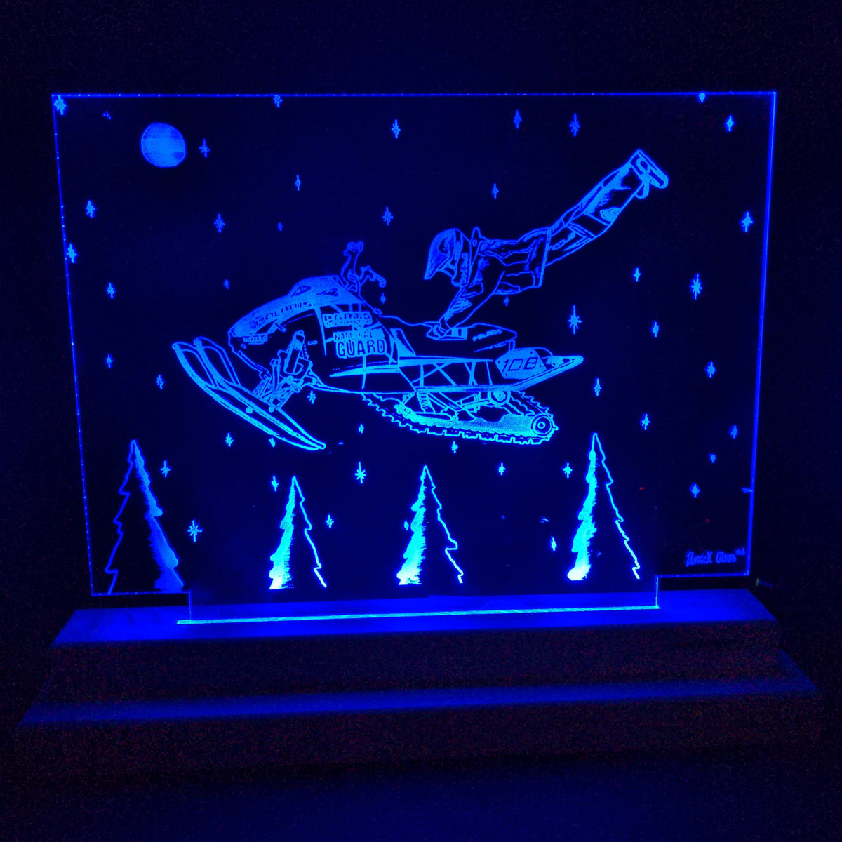 Blue LED-illuminated acrylic panel depicting a U.S. Coast Guard snowmobile with a helmeted figure diving off, above stylized pine trees and stars under a moon.