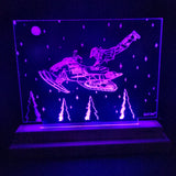 Purple LED-illuminated acrylic panel depicting a person leaping onto a snowmobile above glowing pine trees and stars, with a moon in the top left corner.