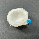 White Stilbite crystal cluster with small sharp formations and a bright blue Cavansite ball attached on a dark gray background.