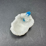 White Stilbite crystal formation with a small vibrant blue Cavansite ball on top, displayed on a dark gray textured surface.