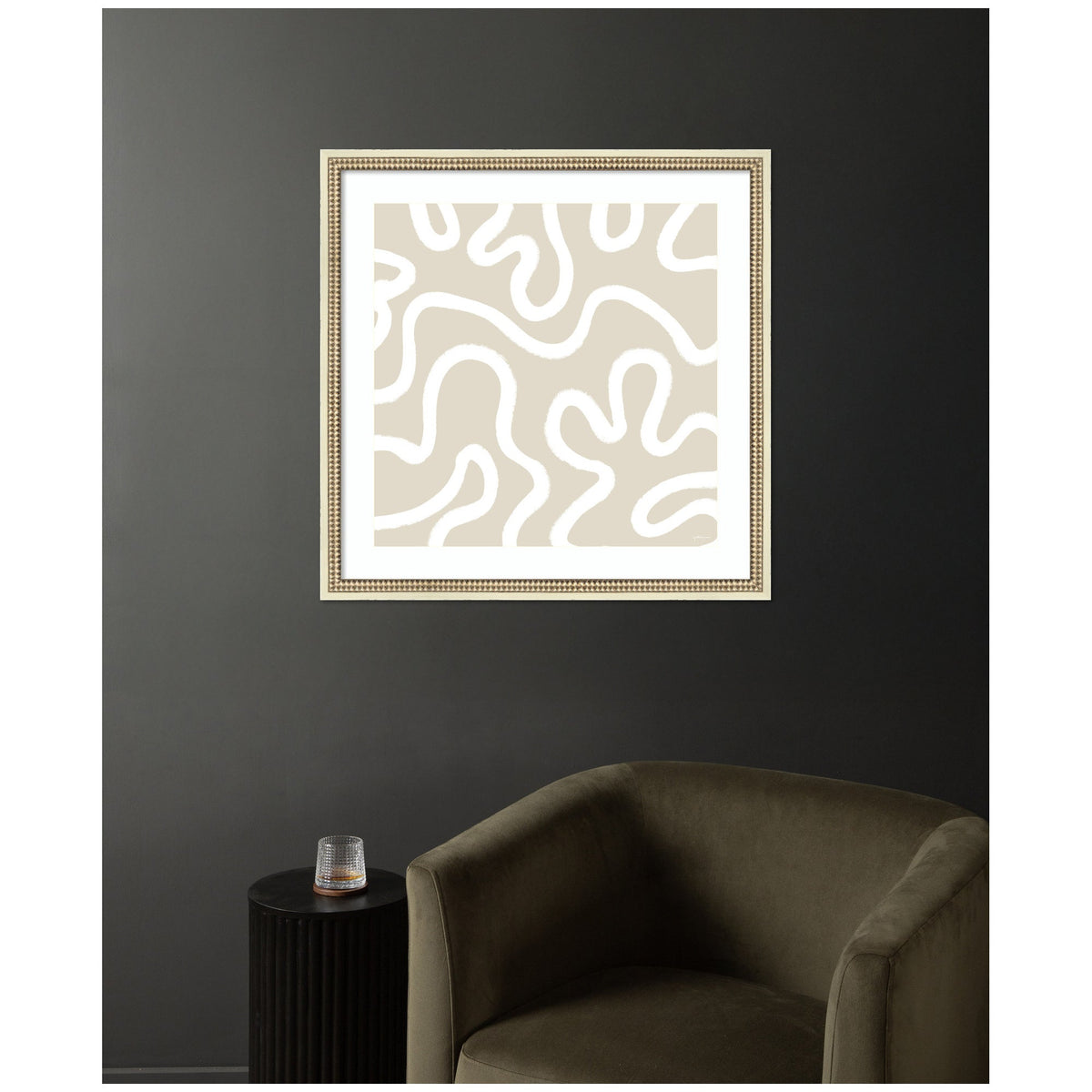 Framed wall art print with beige background and white abstract organic shapes, displayed above an olive green armchair and a black round side table with a glass.