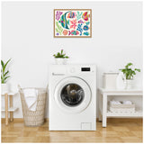 Framed canvas above a white front-load washing machine shows colorful tropical fish in coral reef shapes, set in a clean laundry room with plants and light wood flooring.