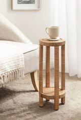 Tall, narrow round side table with woven rattan surfaces and vertical rattan columns, holding a beige ceramic mug, next to a light fabric armchair on a textured carpet.