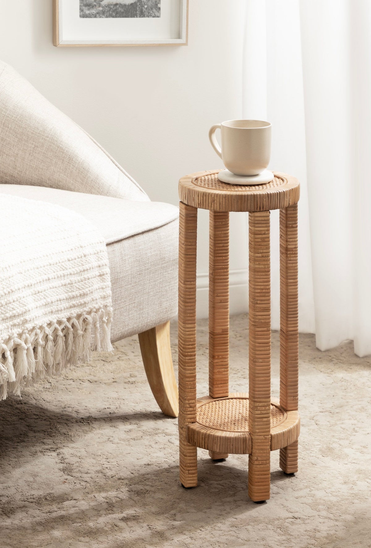 Tall, narrow round side table with woven rattan surfaces and vertical rattan columns, holding a beige ceramic mug, next to a light fabric armchair on a textured carpet.