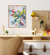 Abstract botanical print with red, yellow, and blue leaves on pale blue background, framed in light wood, hanging above a wooden desk with a beige chair and yellow lamp.
