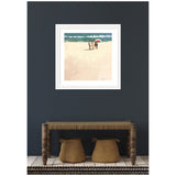 Framed wall art print of three people standing on a sandy beach near ocean waves with a red and white umbrella above them, hung on a dark wall above a wooden bench and two woven baskets.