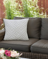 Square white pillow with textured ogee diamond pattern on a dark brown outdoor wicker sofa, set against green plants and wooden fence background.