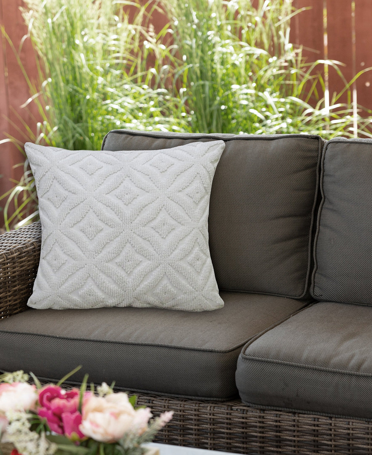 Square white pillow with textured ogee diamond pattern on a dark brown outdoor wicker sofa, set against green plants and wooden fence background.