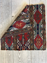 Folded antique tribal cradleface textile featuring geometric patterns in muted red, blue, white, and brown on a light wood floor background.