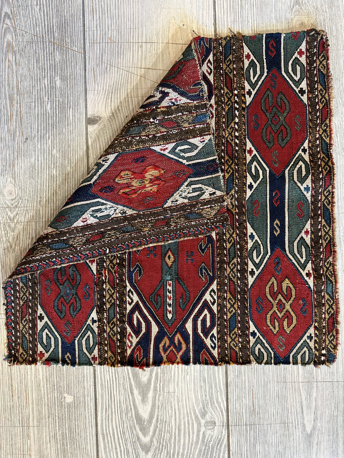 Folded antique tribal cradleface textile featuring geometric patterns in muted red, blue, white, and brown on a light wood floor background.