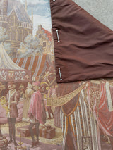 Partial view of a 1990s Belgium wall tapestry depicting medieval market scene with figures in period clothing, striped tents, and a stone building, folded to show brown fabric backing secured with pins.
