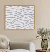 Framed wall art with simple, wavy blue lines on a white background above a beige armchair and a table holding a ribbed translucent vase with foliage.
