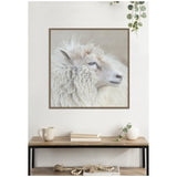 Close-up portrait of a white sheep with thick, curly wool and a calm expression, framed and hanging above a wooden console table with books, a mug, and plants.