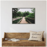 Framed canvas featuring a wooden forest pathway with handrails, surrounded by dense green trees under a cloudy sky, hanging above a wooden bench with a beige welcome home pillow.