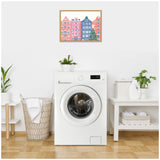 Framed canvas with colorful row of pink and blue townhouses and a decorated Christmas tree hangs above a white front-loading washing machine in a bright laundry room.