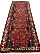 Long vintage Persian Hosseinabad runner rug with a red field featuring geometric red medallions and floral motifs, bordered by a cream band with red and blue floral patterns.