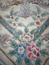 Corner detail of a hand-tufted Chinese Aubusson wool rug featuring blue, pink, and cream floral motifs with green leaves on a beige background framed by teal and brown accents.