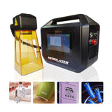 Compact black portable laser engraving machine with a touchscreen and attached orange protective cover, shown with example engravings on metal and plastic surfaces.