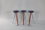 Three modernist bar stools with round dark purple seats, wooden central supports, and angled silver metal legs, arranged side by side on a light grey background.