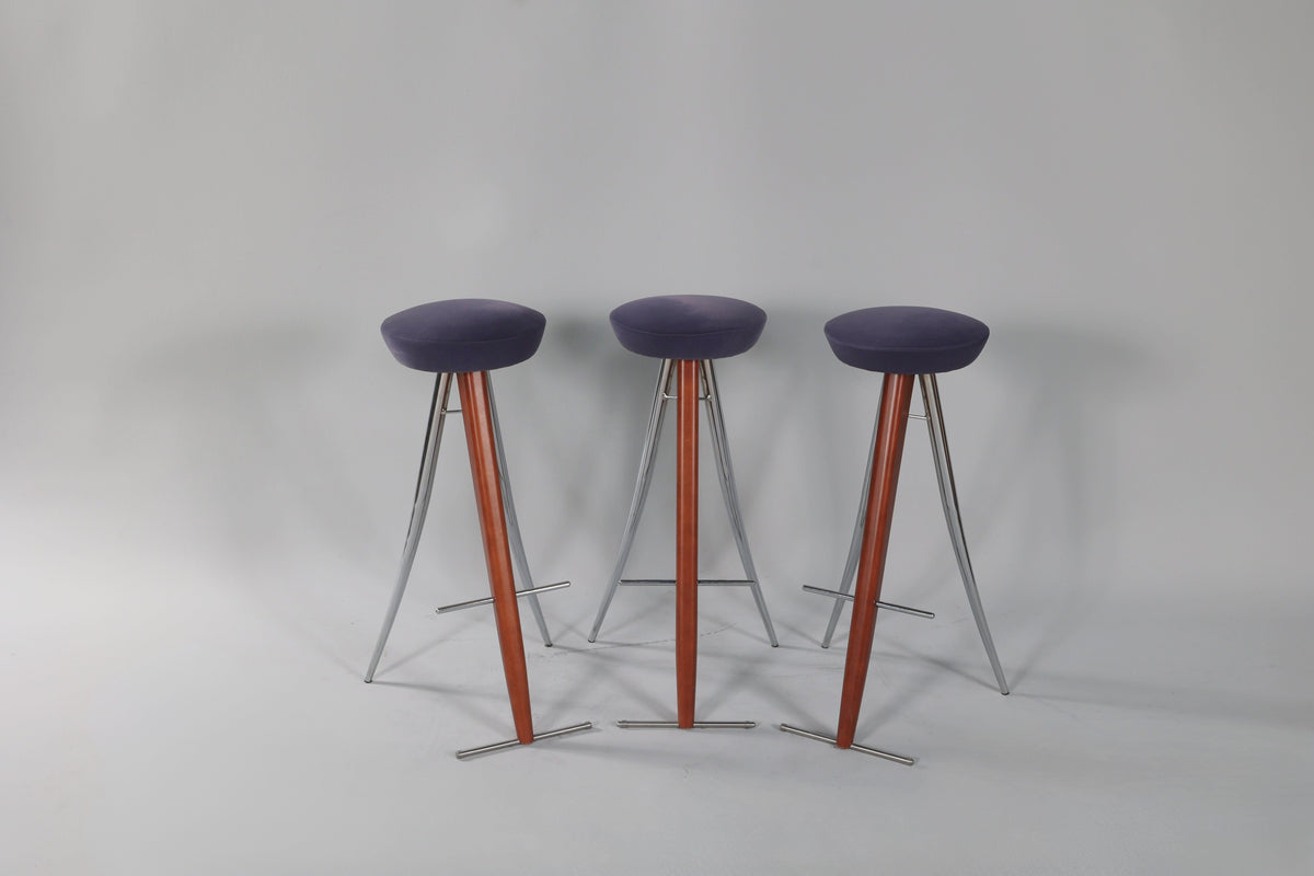 Three modernist bar stools with round dark purple seats, wooden central supports, and angled silver metal legs, arranged side by side on a light grey background.