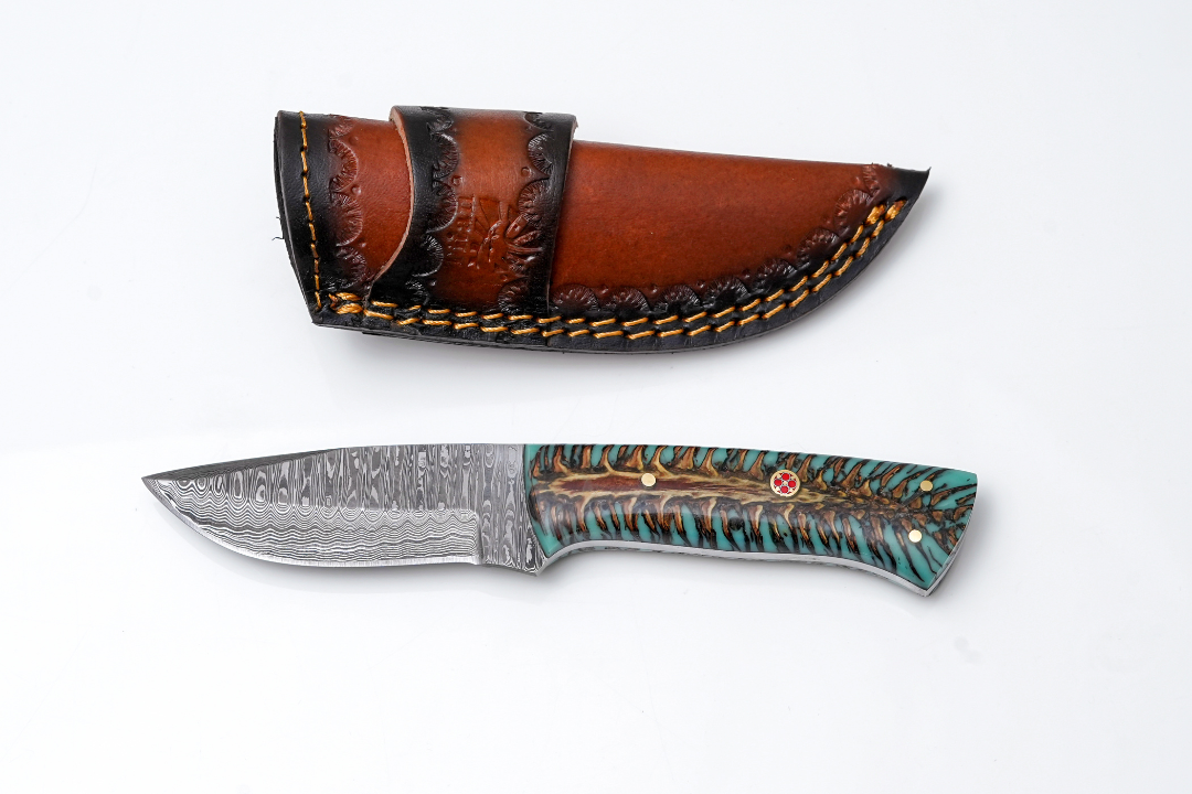 Hand-Forged Damascus Steel Skinner Knife with stabilized pine cone and teal resin handle with mosaic pin next to hand-tooled leather sheath