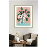 Framed painting of white flowers with green leaves in a blue vase against a blush pink background, hanging above a round wooden table with a white vase and two dark armchairs.