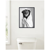 Black and white framed canvas of a lioness wrapped in a white towel robe, hung on a white bathroom wall above a toilet with a silver flush handle.