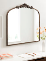 Arched wall mirror with a dark wooden frame and decorative carved detail at the top center, reflecting a bright window and simple room interior.