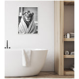 Black and white framed canvas showing a lion wrapped in a towel robe, hung above a white freestanding bathtub in a minimalist bathroom with wooden shelving.
