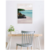 Framed canvas print of a coastal scene with rocky shore, green foliage, and calm blue water hanging above a modern wooden desk with plant and metal decor, against a white wall.