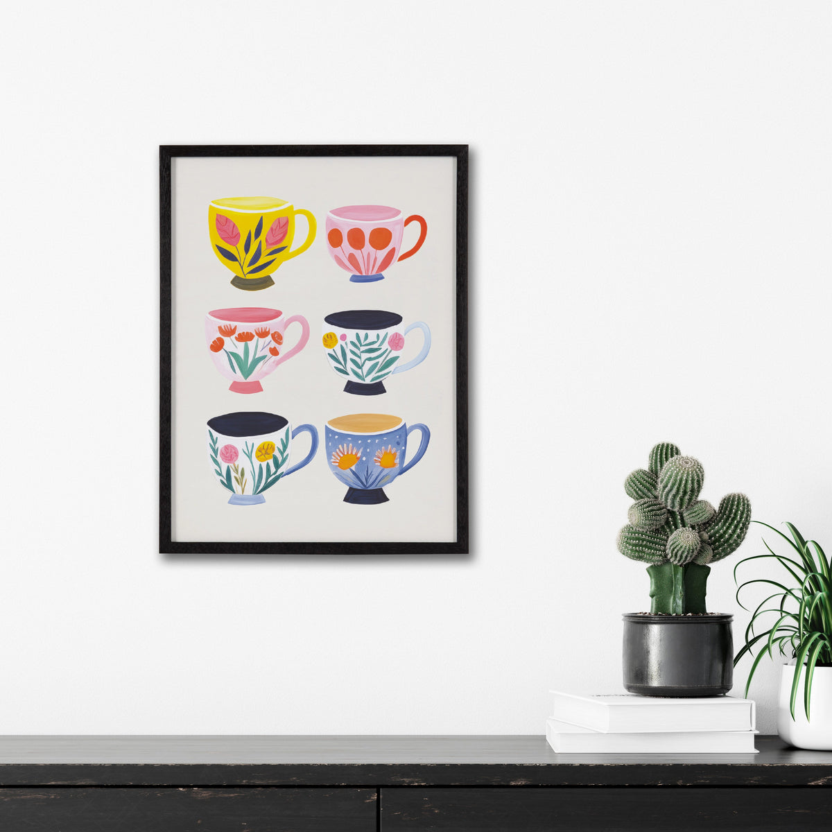 Framed print featuring six colorful tea cups with distinct floral designs in three rows, hung on a white wall above a black surface with potted plants and books.