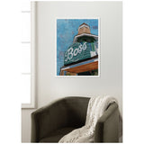 Framed canvas print of a vintage green and white sign reading 'The Boss' against a clear blue sky, hanging above a dark chair with a knitted blanket.