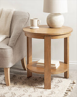 Round wood side table with a lower shelf holding stacked books, topped with a cream ceramic lamp and a matching coffee cup, next to a beige upholstered armchair.