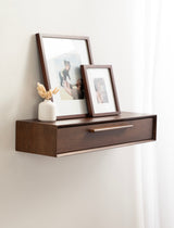 Wall-mounted dark wood floating console table with a drawer, topped with two framed photos and a small white vase holding dried beige flowers, against a light wall and sheer white curtain.
