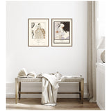 Two framed vintage-style canvas art prints with portrait illustrations, hung side by side above a wooden bench draped with a light fabric in a minimalist light-filled room.