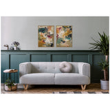 Two framed abstract paintings featuring warm beige, yellow, teal, and burgundy brushstrokes hang side by side on a light gray wall above a modern light gray sofa.