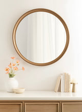 Round wall mirror with a thin wooden frame reflecting sheer white curtains, above a light wood cabinet decorated with a white vase of peach flowers, books, and a candle holder.