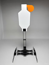 White metal shooting target shaped like a human silhouette with a small bright orange circular area on the upper right, mounted on a black adjustable metal stand with angled feet.