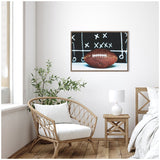 Framed canvas shows a brown football with white laces in front of a black chalkboard playbook featuring white Xs and Os, hung on a white wall above a wicker chair and wooden table.