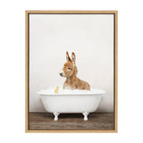 Young brown donkey sitting in a white clawfoot bathtub filled with bubbles, accompanied by a small yellow rubber duck on a wooden floor background.