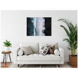 Framed aerial photograph of foamy ocean waves crashing onto a dark black sand beach, hung above a white sofa with patterned cushions and flanked by green plants.