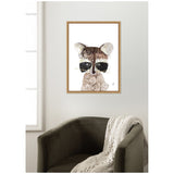 Framed canvas art print of a stylized raccoon portrait with dark eye mask and beige-brown fur, hanging on a wall above a dark green armchair draped with a cream knit blanket.