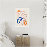 Framed abstract print with pink wavy lines, a bold blue irregular shape, and two orange rounded forms hanging above a rustic wooden bedside table with books and a cup.